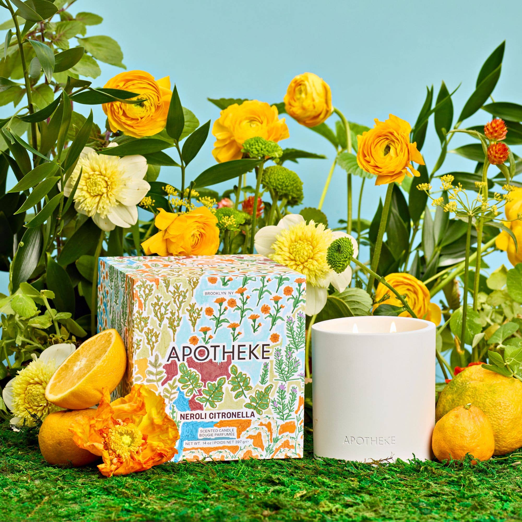 Apotheke Neroli Citronella candle and packaging surrounded by flowers and fruits on a grassy background